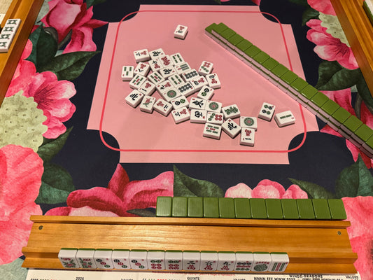 Mahjong game setup with white tiles featuring black and red Chinese characters, arranged on a pink playing field over a floral mat with pink flowers and green leaves, plus wooden racks holding green-backed tile backs.