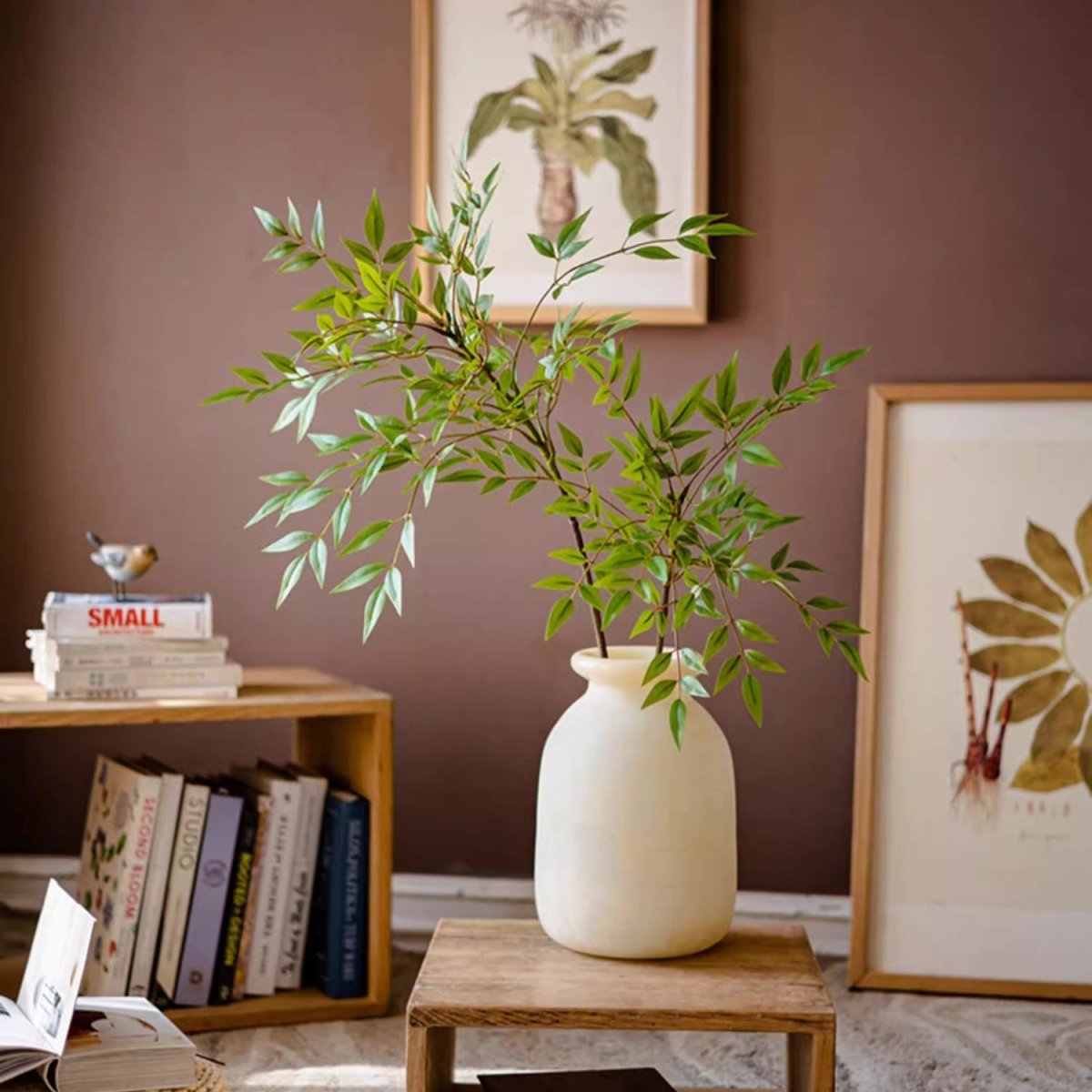 White vase with green plant on a wooden table against a brown wall with framed artwork.