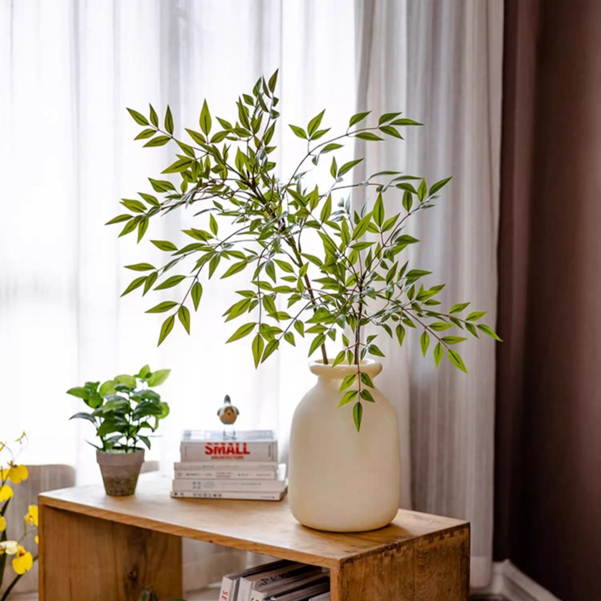 Decorative setup with a vase of greenery on a wooden table against a white curtain background