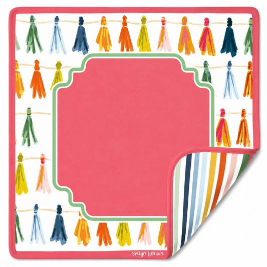 A square mahjong mat  coral pink center label area, bordered by colorful hanging tassels on a white background. One corner is folded over to show a striped reverse side with blue, green, orange, yellow, and white stripes