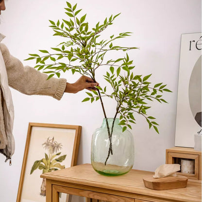 Person arranging green branches in a clear glass vase on a wooden table.